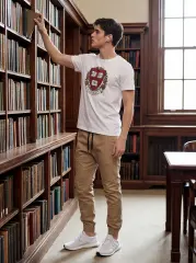 A male model in a white t-shirt and the khaki jogger pants, standing in a library looking at a book. The setting shows the joggers in a smart-casual, everyday environment.