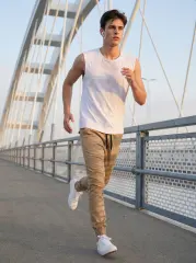 A full-body view of a male model running across a white bridge, wearing the khaki jogger pants. The scene emphasizes movement and active lifestyle, paired with a white sleeveless top and white sneakers.
