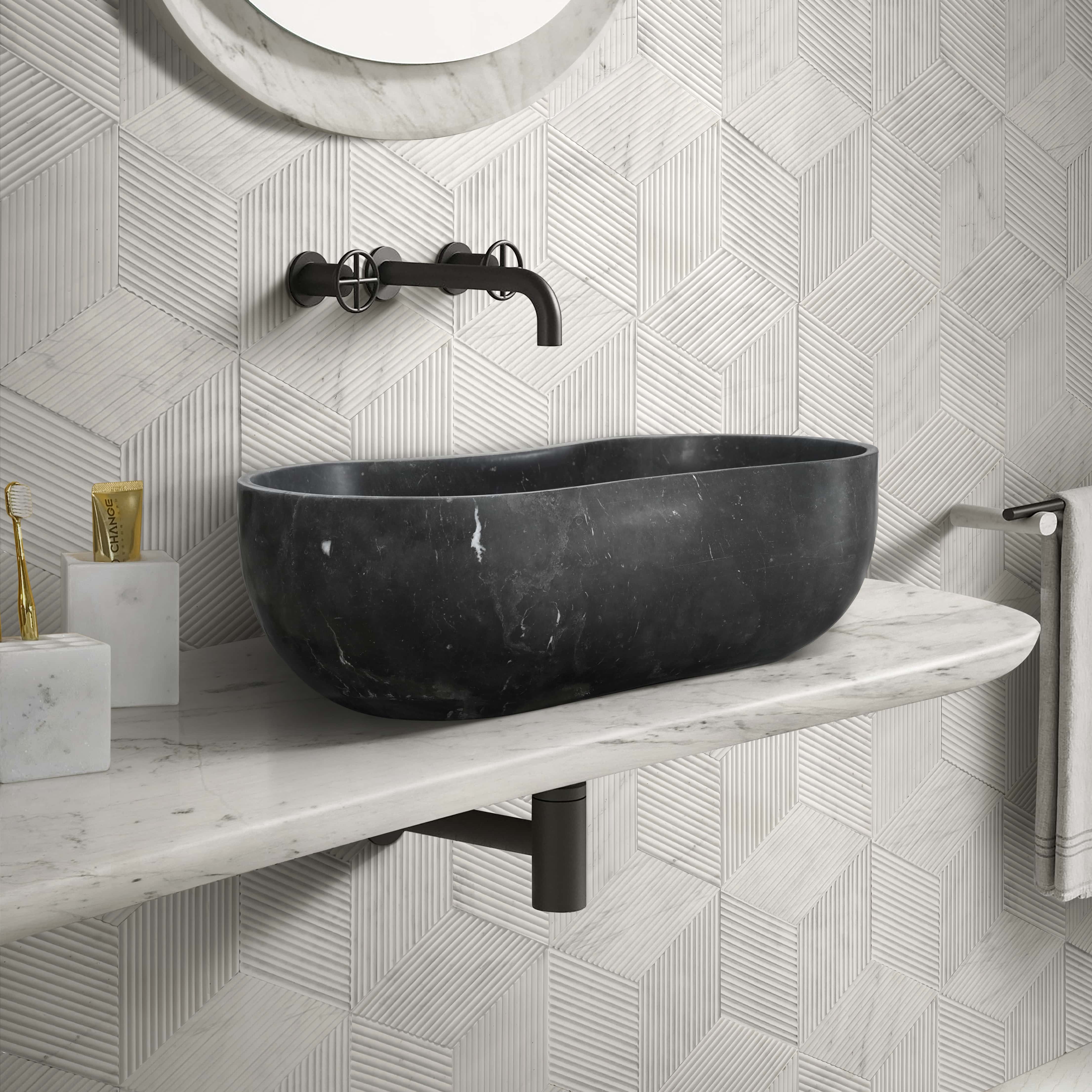 A honed Olive Black natural marble wave vessel sink and Snow Mountain White marble accessories against a Snow Mountain White hexagon OL textured finished tile feature wall.
