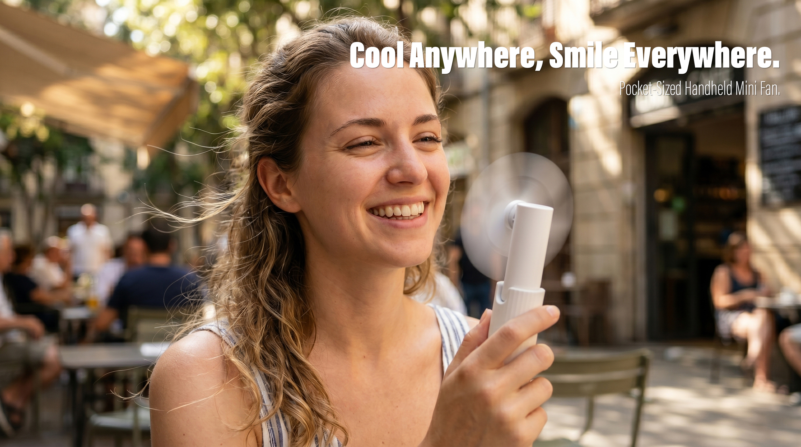 A close-up lifestyle shot of a smiling young woman holding a white minimalist handheld mini fan with spinning blades while walking past outdoor cafes on a sunny street.