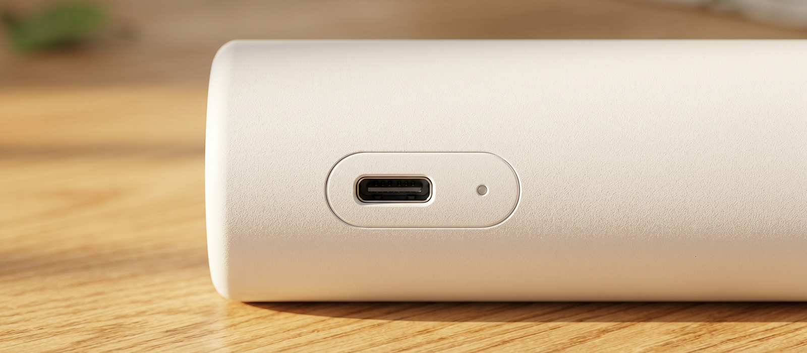 A clean macro photograph of the Type-C charging input port and precision recessed pinhole LED indicator light on the base of a white minimalist portable table lamp resting on a sunlit wooden desk