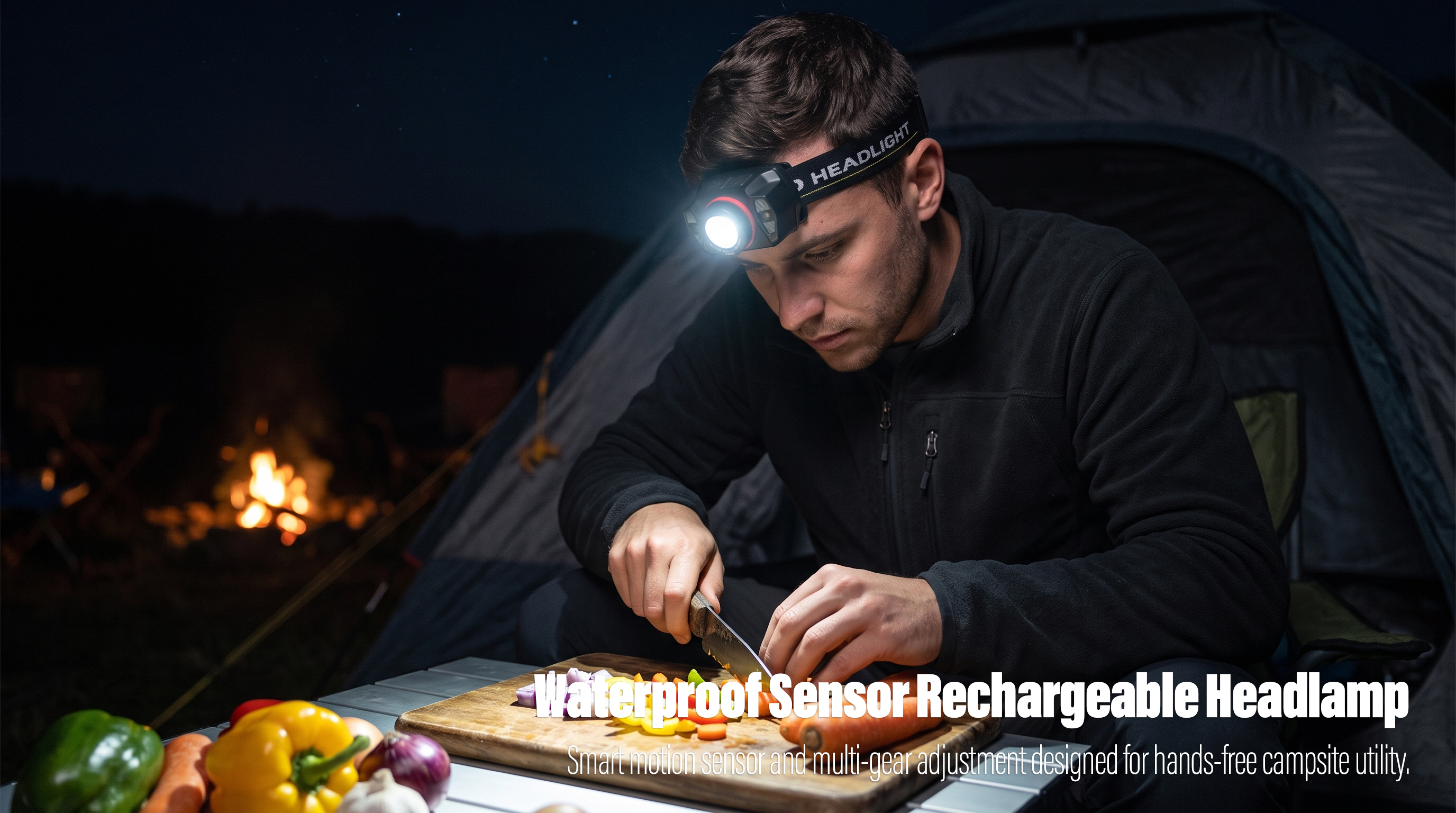 A real-world outdoor lifestyle photo of a man sitting at a folding table at night, using a knife to slice carrots and peppers on a wooden cutting board, while his black rechargeable sensor headlamp casts a bright beam onto the ingredients, with a tent and a campfire in the background.