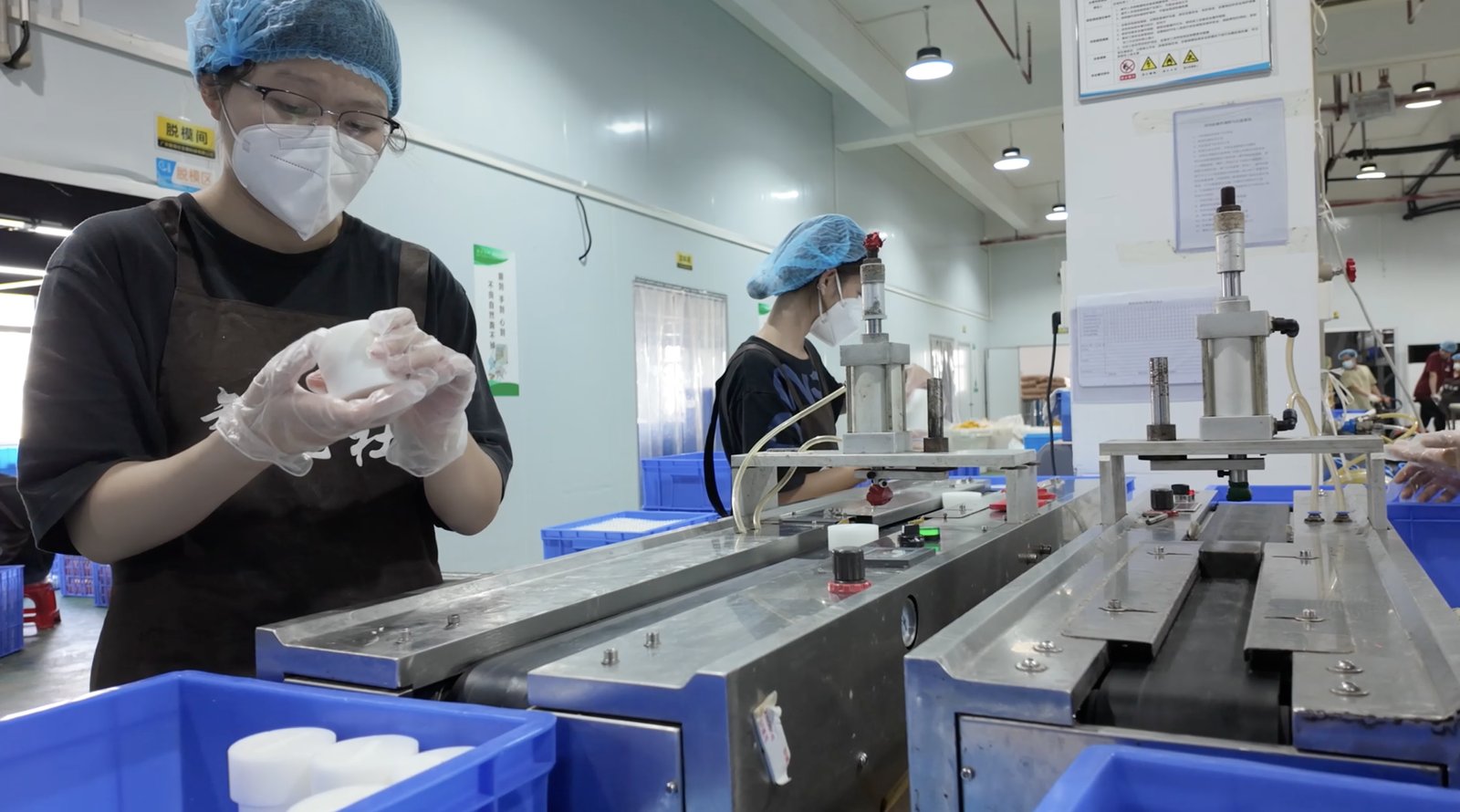 A worker wearing gloves, a mask, and a hairnet inspecting a white soap bar near a production line in a factory.