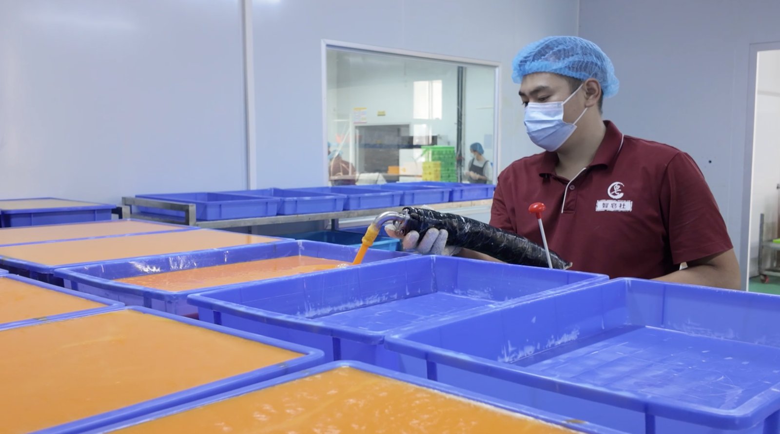 A worker wearing a mask and hairnet pouring liquid into blue trays filled with orange-colored soap mixture in a clean production facility.