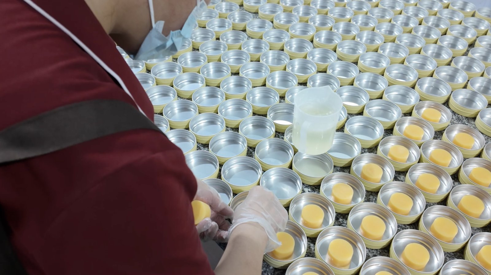 A worker wearing gloves and a mask placing a round soap into a metal container, with multiple containers arranged on a table.