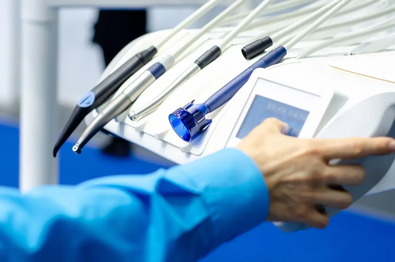 Dentist's hand in a blue shirt adjusting the control panel next to a tray of dental handpieces.