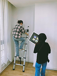 две девушки вешают картины с помощью одной кнопки выдвижной лестницы