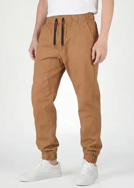 Close-up front view of ItalyMorn men's light brown jogger pants on a model, highlighting the comfortable elastic waistband with black drawstrings, deep side pockets, and modern slim-tapered fit. Paired with white sneakers
