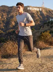 A young man trail running in the hills with the Hollywood sign in the background, wearing a light grey t-shirt and ItalyMorn men's dark grey jogger pants, demonstrating the pants' high flexibility for athletic performance