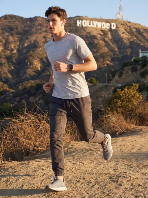 A young man trail running in the hills with the Hollywood sign in the background, wearing a light grey t-shirt and ItalyMorn men's dark grey jogger pants, demonstrating the pants' high flexibility for athletic performance