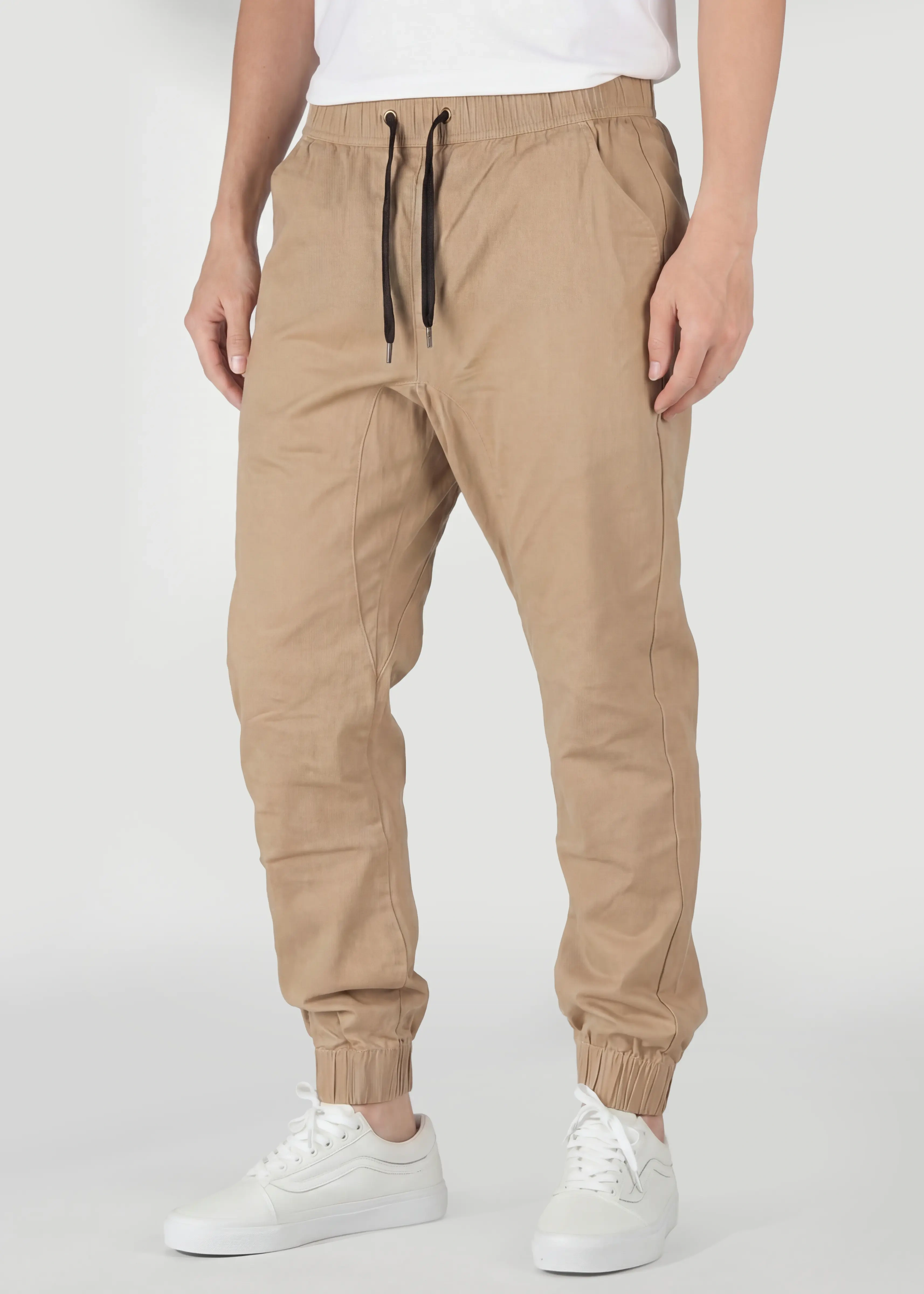 A detailed close-up view from the front of ItalyMorn men's khaki jogger pants, showcasing the elastic waistband with drawstrings, practical front pockets, and ribbed cuffs. A model's lower body is shown, wearing white sneakers.