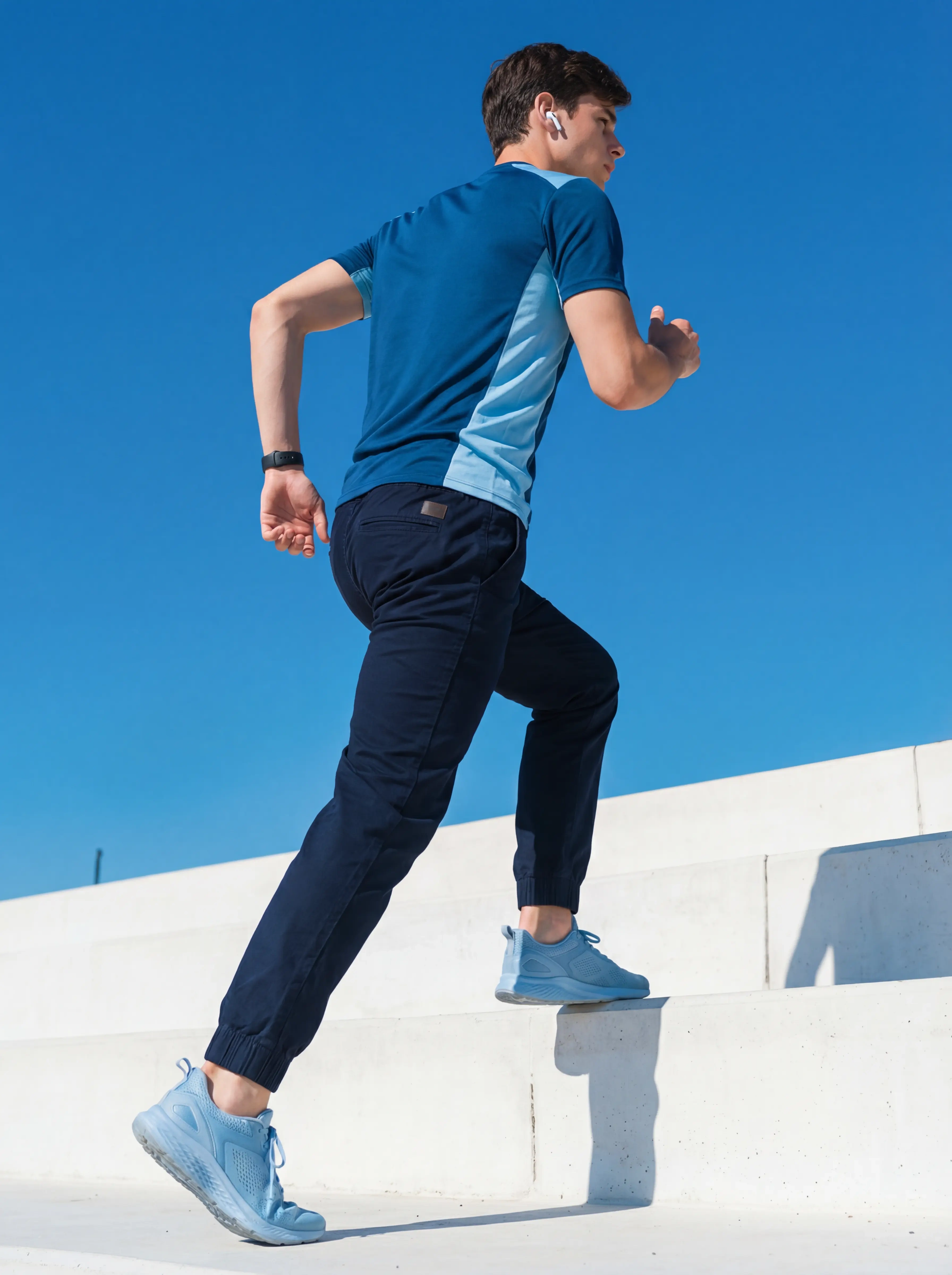  A man running up outdoor white stairs against a clear blue sky wearing ItalyMorn men's navy blue jogger pants, demonstrating the fabric's flexibility and breathability for athletic activities
