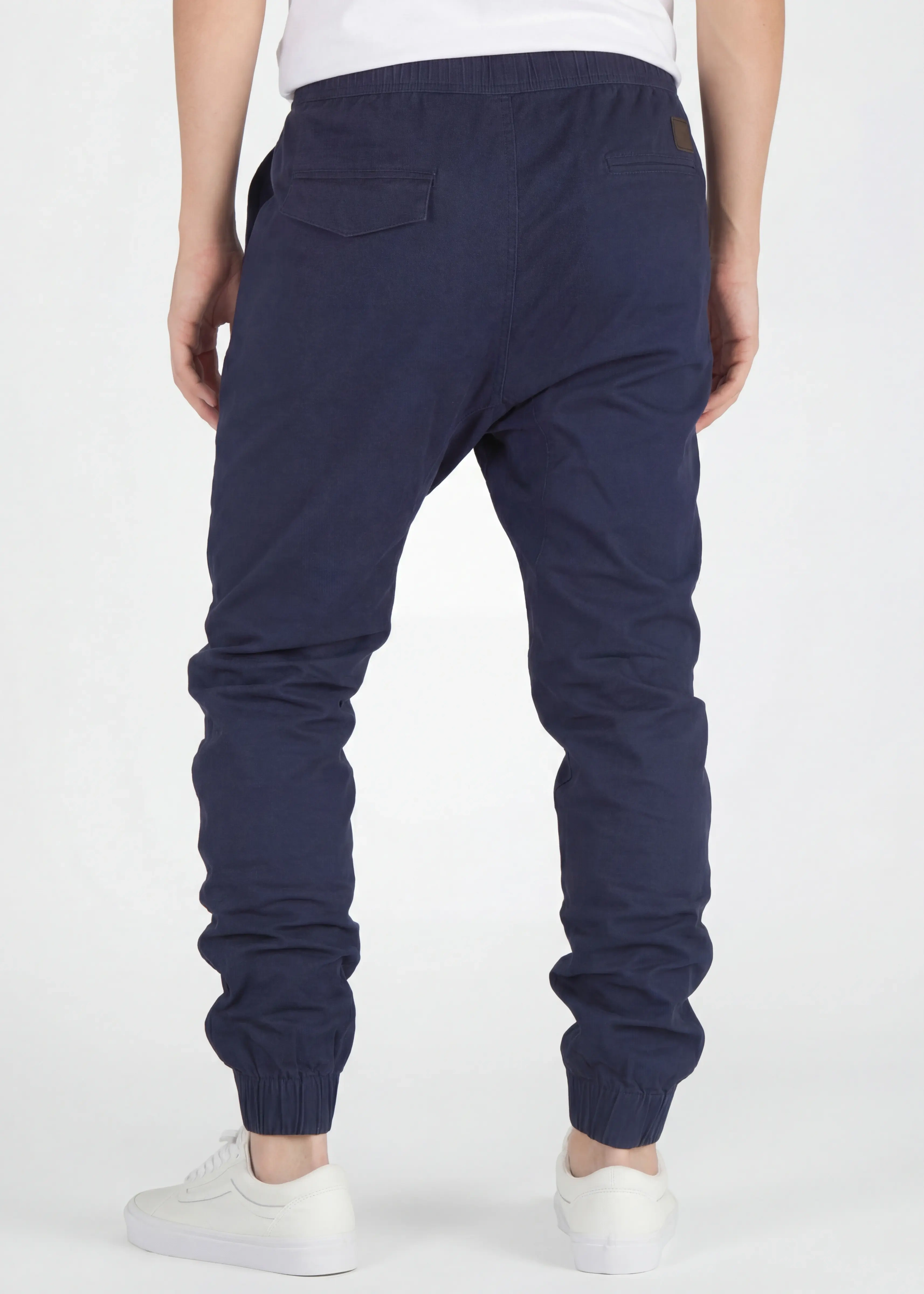 A rear close-up view of ItalyMorn men's navy blue jogger pants, showcasing the clean tailored fit, functional back pockets with flap detail, and secure ribbed ankle cuffs. Paired with white sneakers.