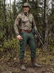 A hunter in a camouflage shirt and rubber boots standing in the woods holding a rifle, utilizing the deep side pockets of his ItalyMorn men's army green cargo pants for gear. This highlights the pants' tactical and field utility.