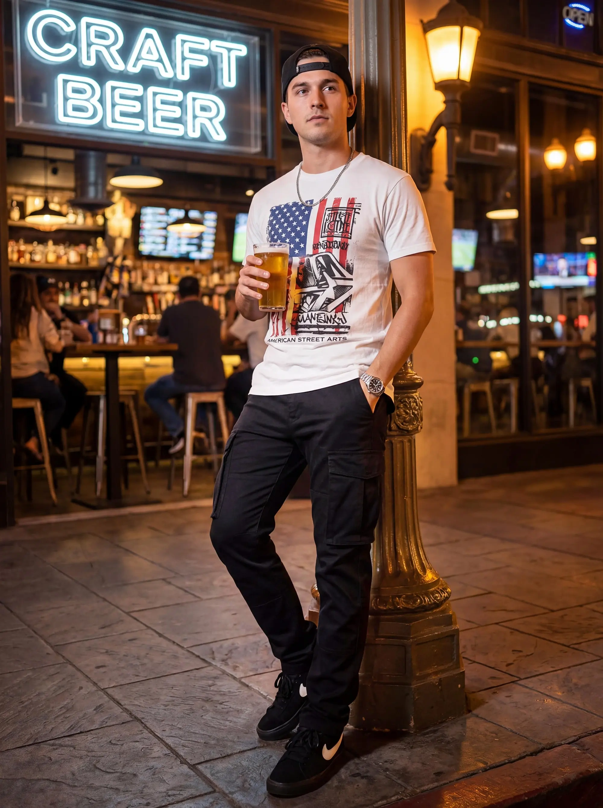 A stylish young man leaning against a lamppost outside a craft beer bar at night, wearing a graphic t-shirt and ItalyMorn men's black cargo pants, demonstrating a relaxed, casual urban outfit.