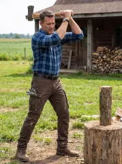 A robust man swinging an axe to chop wood outside a rustic cabin, wearing a blue plaid shirt and ItalyMorn men's coffee brown cargo pants with work gloves tucked into the pocket, emphasizing rugged workwear utility