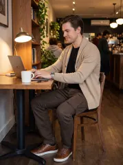 A man working on a laptop at a cozy cafe table, wearing a beige cardigan, brown leather shoes, and ItalyMorn men's coffee brown cargo pants. This highlights the pants' versatility for a smart-casual or remote work lifestyle.