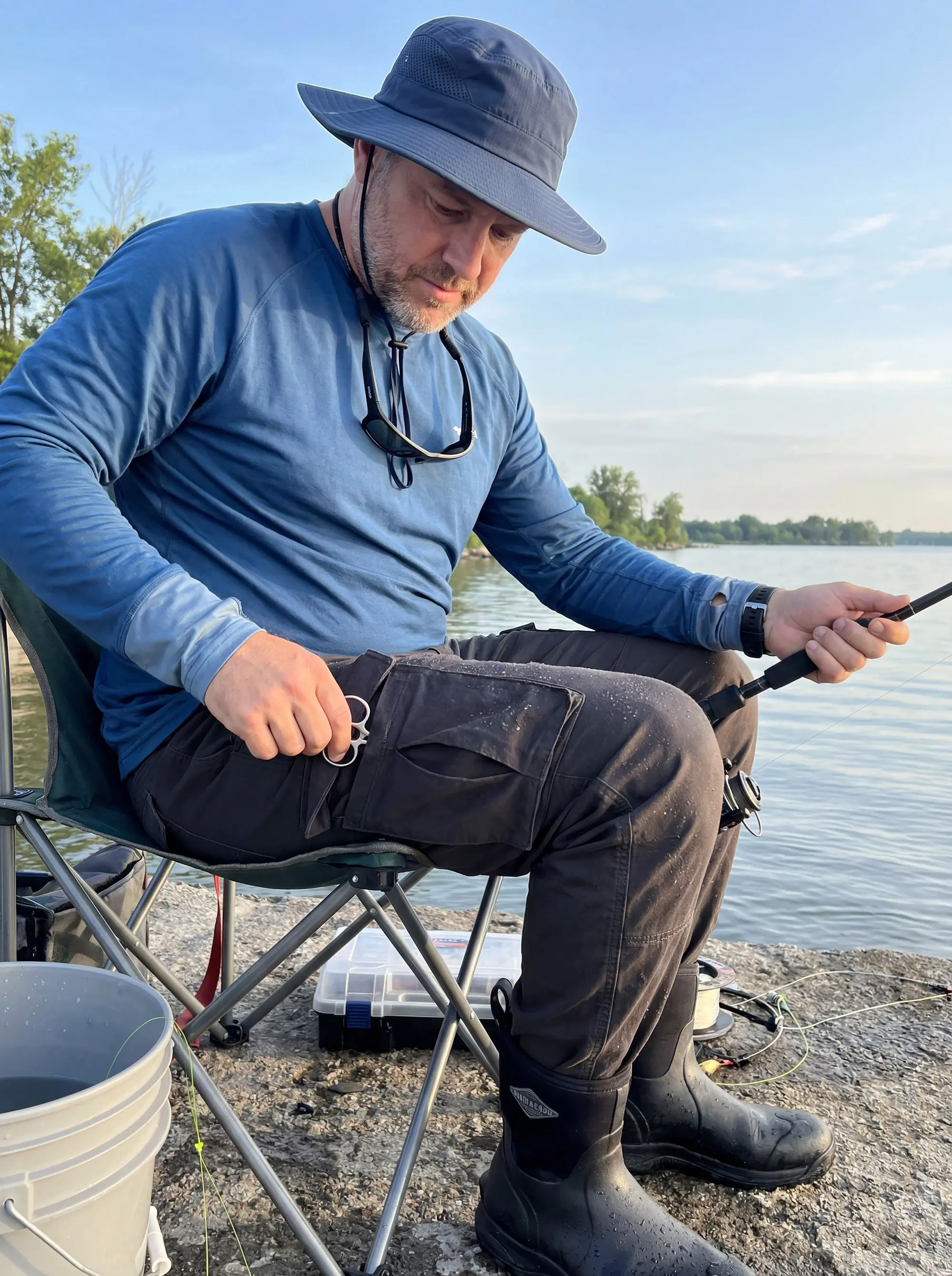 A man sitting by a lake fishing, easily accessing a tool from the functional side cargo pocket of his ItalyMorn men's dark grey cargo pants, showcasing their durability and convenience for outdoor hobbies.
