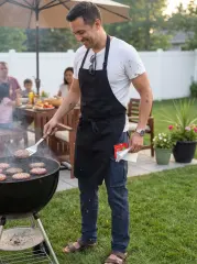 A smiling man grilling burgers at a backyard family BBQ, wearing an apron, white t-shirt, and ItalyMorn men's navy blue cargo pants. This showcases a relaxed and comfortable outfit for casual weekend gatherings.