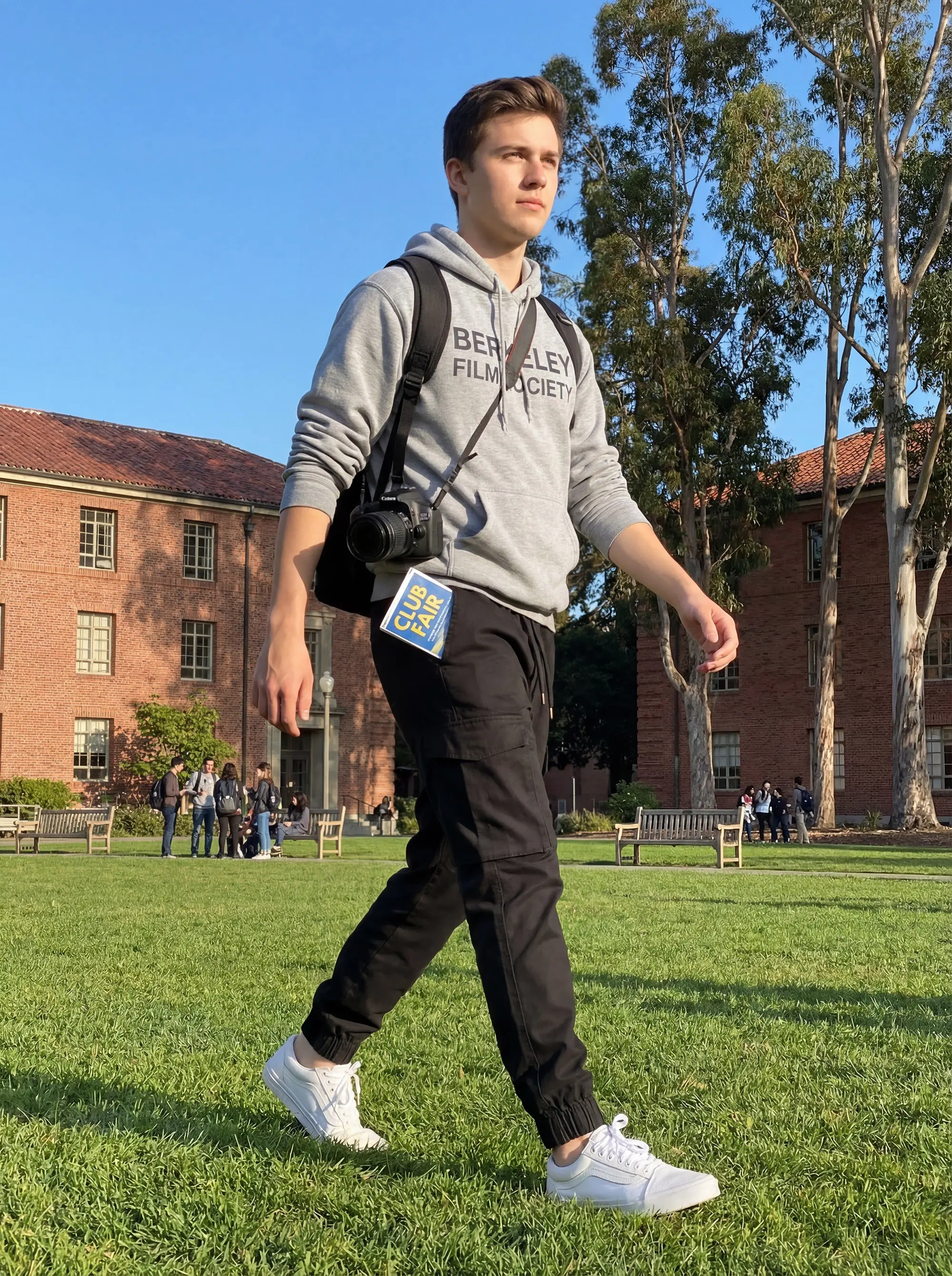 A young man walking confidently on a sunny college campus, wearing a grey hoodie and men's black cargo joggers with white sneakers, carrying a camera and a backpack.