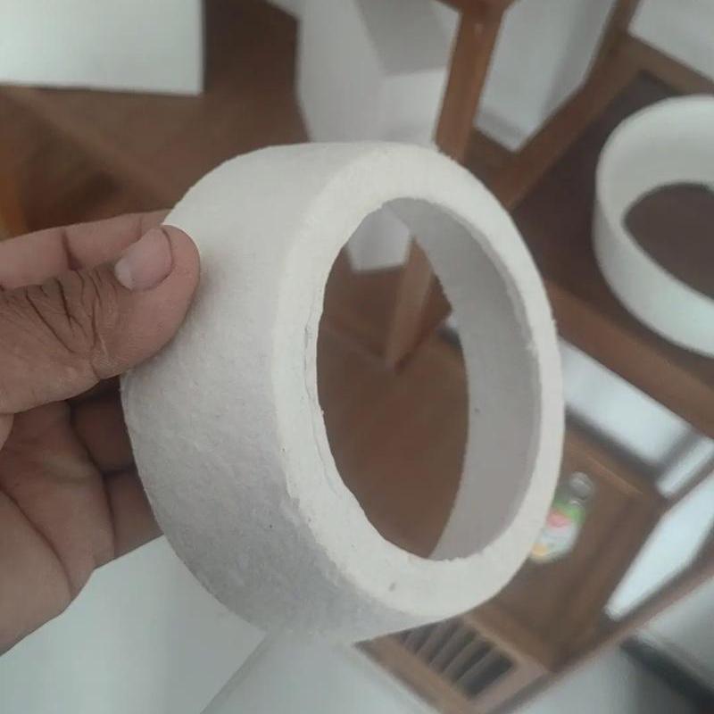A close-up view of a hand holding a customized, circular white ceramic fiber insulating ring, showing its thickness and annular structure detail in front of a wooden display shelf——LiLuan Technology