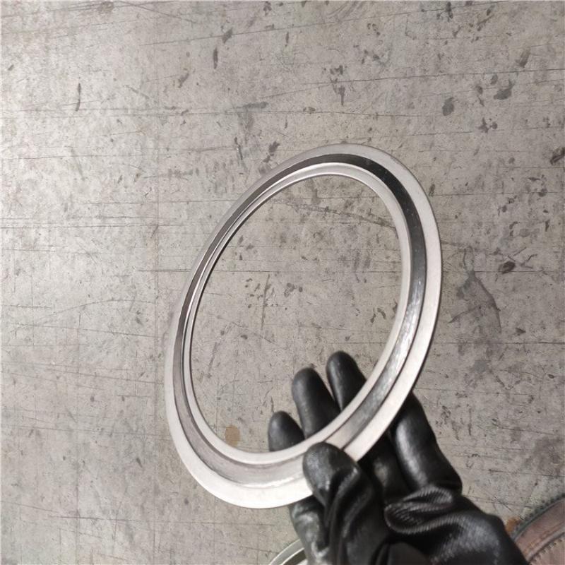 A perspective photograph showing a hand wearing a black industrial glove holding a single armored stainless steel coated flexible graphite gasket. The gasket is held at an angle against a concrete industrial floor background, demonstrating its slim profile and ease of handling for installation into pipe flange sealing systems.---LiLuan Technology