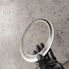 A perspective photograph showing a hand wearing a black industrial glove holding a single armored stainless steel coated flexible graphite gasket. The gasket is held at an angle against a concrete industrial floor background, demonstrating its slim profile and ease of handling for installation into pipe flange sealing systems.---LiLuan Technology