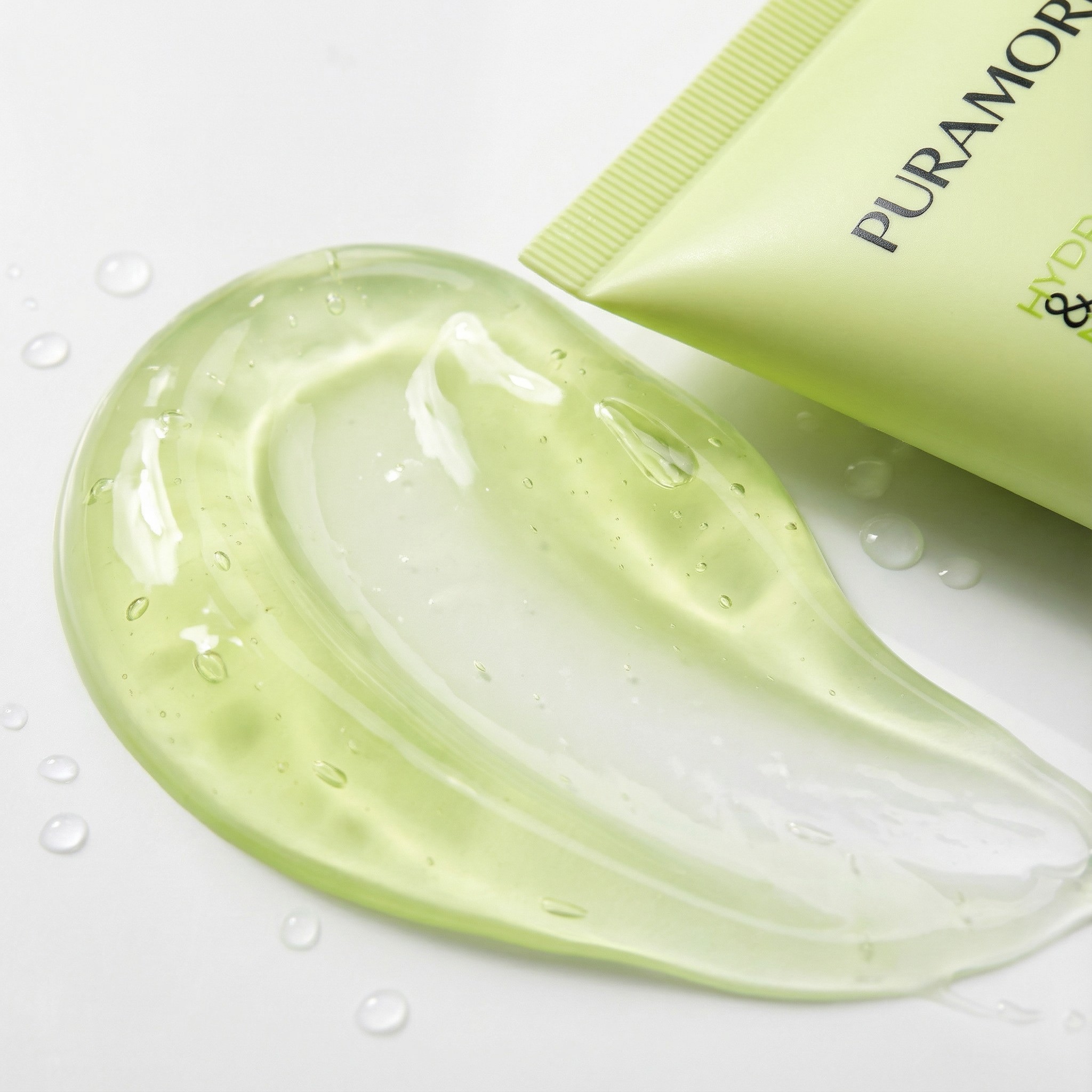Macro close-up texture shot of PURAMORIA Gentle Cleanser gel, displaying a clear, refreshing swipe with realistic water droplets on white background. Illustrates a non-stripping, hydrating facial cleanser texture.
