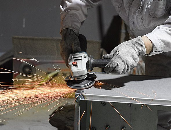 an industrial worker using an angle grinder with a FUJI JITAN wheel on a large stainless steel structure in a factory setting