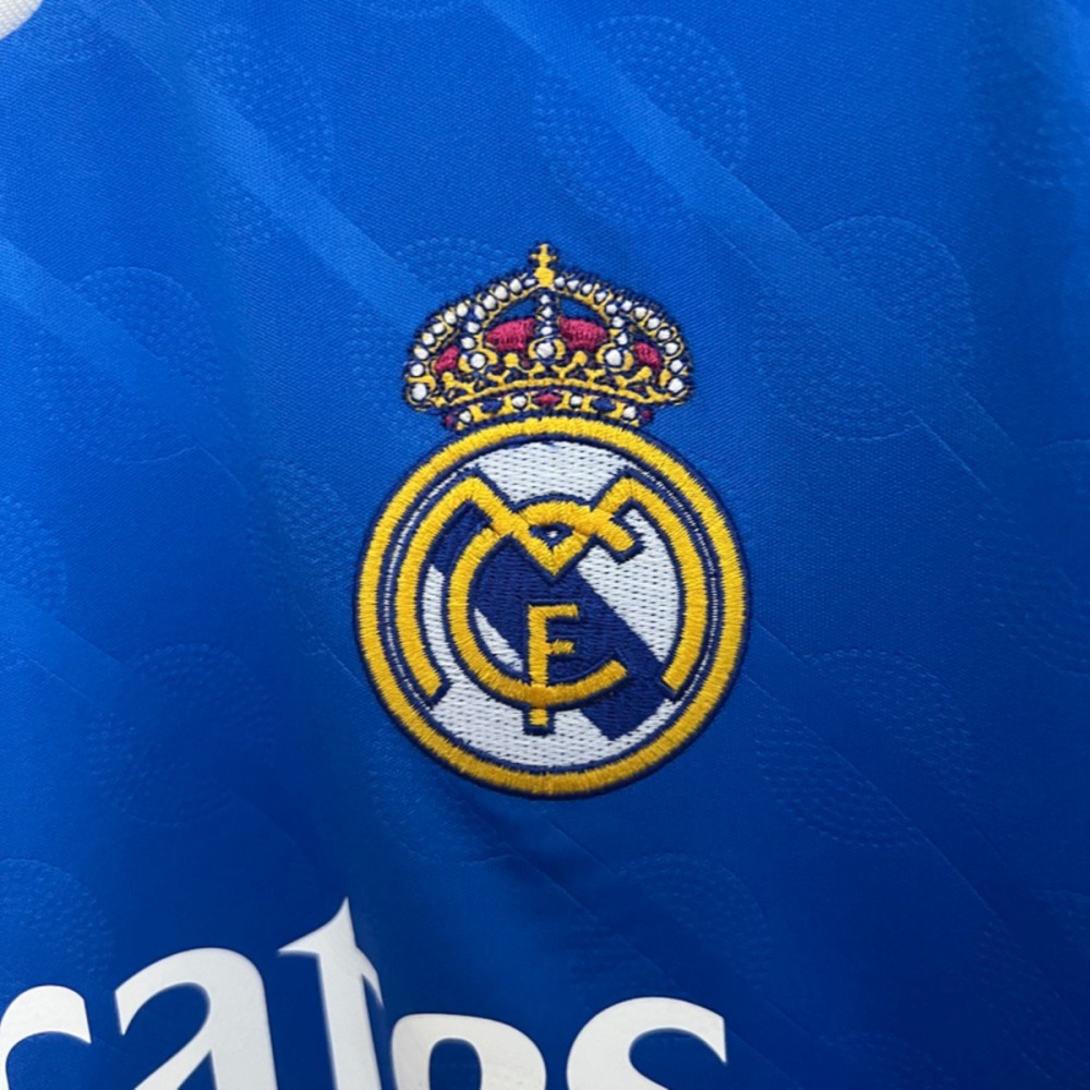 Close-up of embroidered Real Madrid club crest on the 2025-26 second away jersey fan version, highlighting detailed stitching and blue fabric texture.
