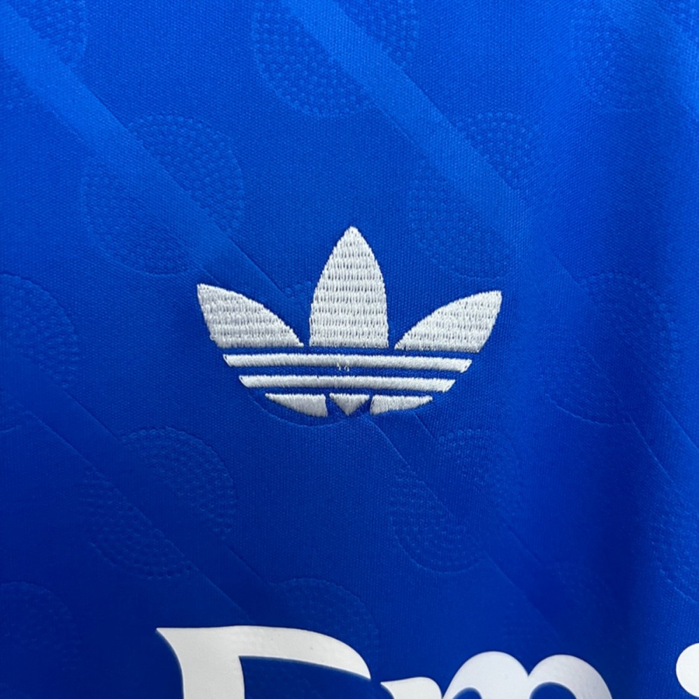 Close-up detail of the 2025-26 Real Madrid second away fan version jersey showing white Adidas Trefoil logo on blue textured fabric.