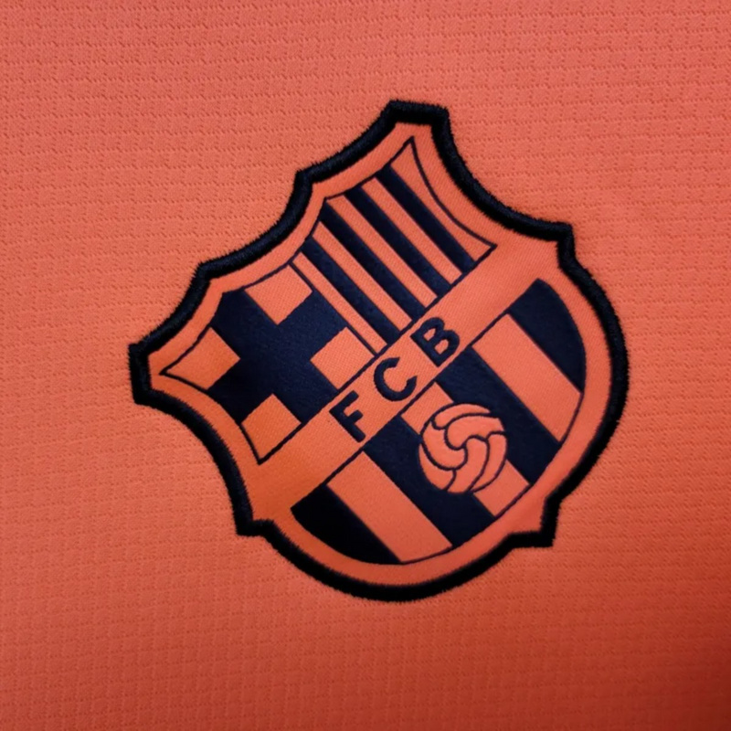 Close-up of the FC Barcelona embroidered crest on the 2025-26 second away jersey, dark blue logo on orange background with detailed fabric texture.
