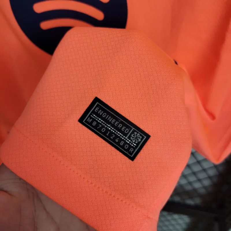 Close-up of the 2025-26 FC Barcelona Second Away Jersey authenticity label showing ENGINEERED tag and product code on textured orange fabric.