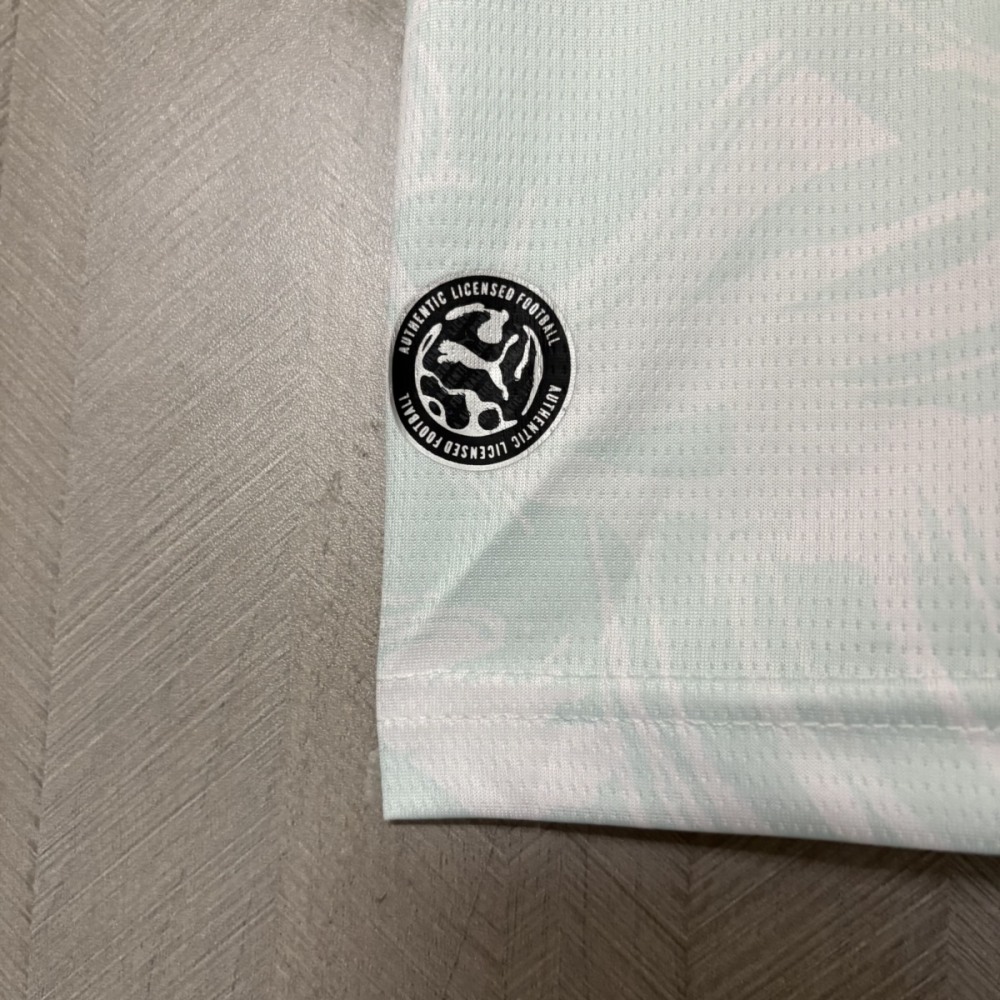 Close-up of the lower hem of the Portugal 2026 World Cup away jersey fan version showing the black circular Puma “Authentic Licensed Football” badge on a white fabric with subtle teal wave graphics.