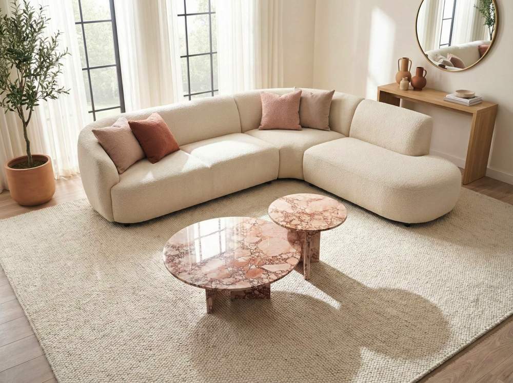 Sun-drenched living room setting featuring a modern curved sofa, an indoor olive tree, and luxury round pink marble nesting tables. The sunlight beautifully reflects off the polished, unique natural stone surface.
