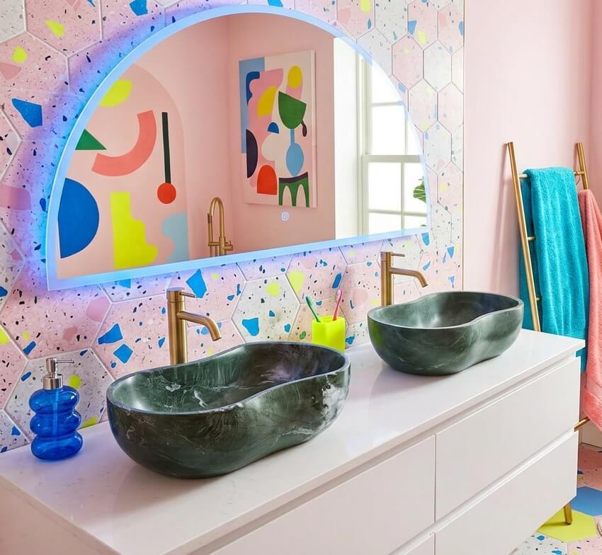 Double Diamond Green marble sinks on a white modern vanity with a hexagonal colorful terrazzo backsplash, featuring abstract pop art and an arched backlit mirror.
