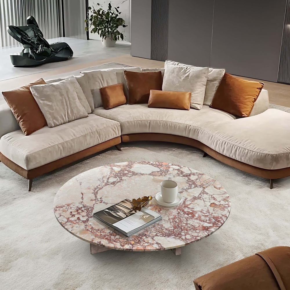A round Diamond Pink marble coffee table with a polished surface, styled in a contemporary lounge featuring a curved sectional sofa and sculptural art.
