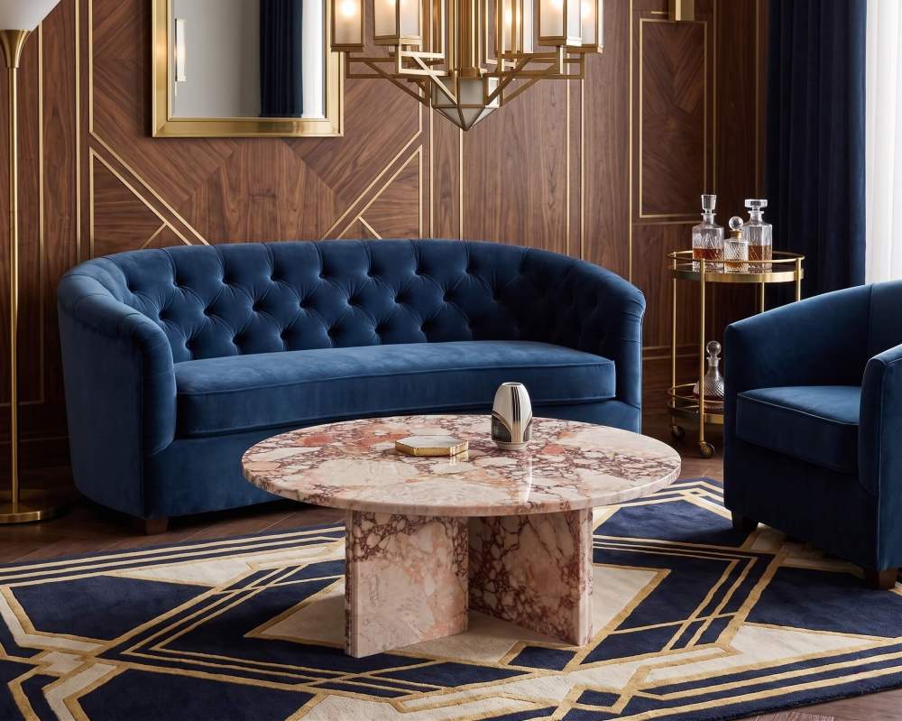 Diamond Pink marble coffee table centered in an Art Deco living room — navy button-tufted velvet sofa, walnut wood-paneled wall with brass geometric inlay, geometric brass chandelier, gold patterned navy area rug, crystal decanters on brass bar cart.