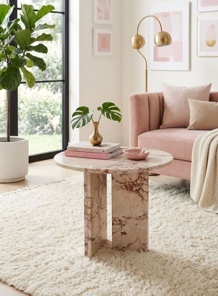 Diamond Pink marble round coffee table styled in a blush and gold feminine living room — pink velvet sofa, gold arc floor lamp, monstera leaves in gold vase on stacked books, shaggy ivory rug and fiddle leaf fig plant.