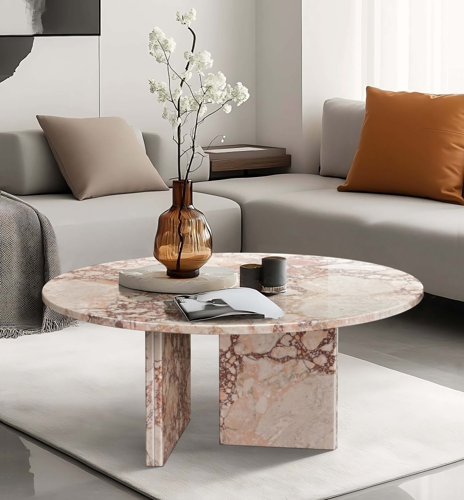Diamond Pink marble round coffee table in a contemporary living room — light gray sectional sofa with camel accent pillow, herringbone throw, abstract monochrome wall art, amber ribbed glass vase with white cherry blossom branches on marble surface, diffused natural light from sheer curtains.
