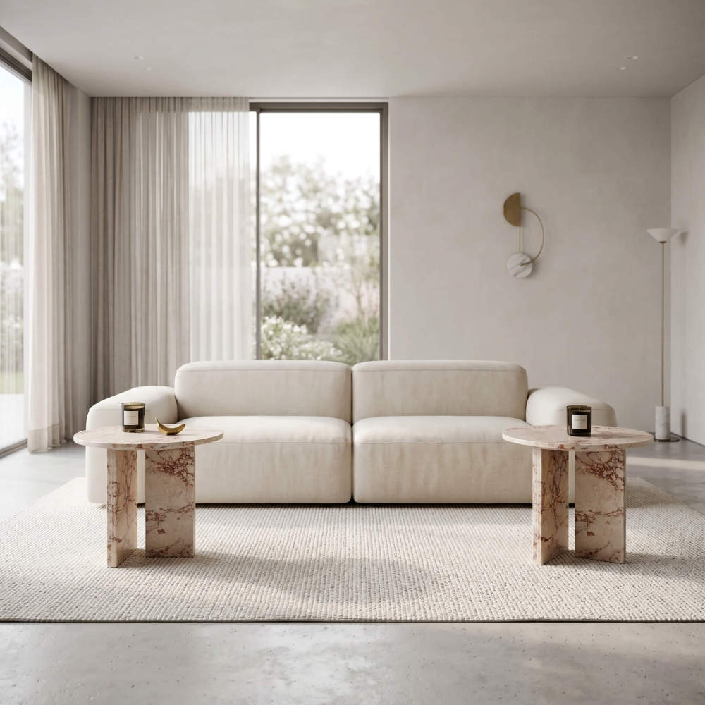 Two Diamond Pink marble side tables placed symmetrically on either side of a cream modular sofa in an all-white contemporary minimalist living room — polished concrete floor, sheer floor-to ceiling curtains, gold half-circle wall art.