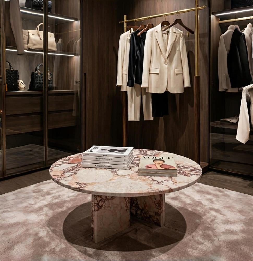 Diamond Pink marble round coffee table as a centerpiece in a luxury women's dressing room — dark walnut cabinetry with glass display cases showing designer handbags, brass clothing rail with tailored blazers, fashion magazines including Vogue stacked on polished marble surface, rose-gray silk-feel area rug.