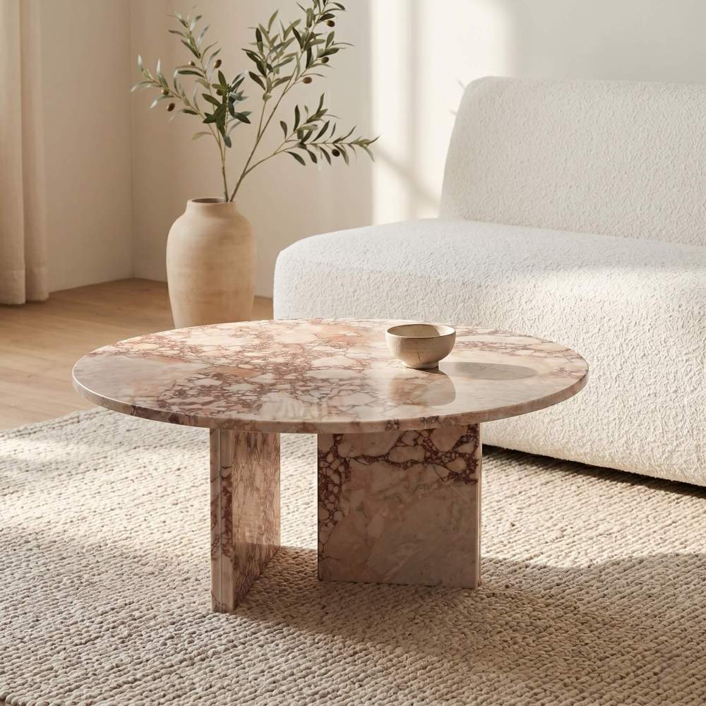 Diamond Pink marble round coffee table in a wabi-sabi minimalist living room — cream bouclé sofa, chunky knit area rug, terracotta vase with olive branches, single ceramic bowl on marble surface, raking morning light casting geometric shadow on warm white wall.