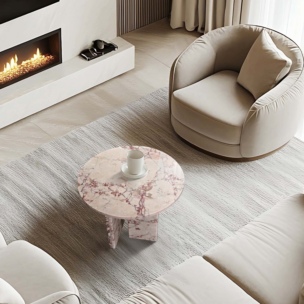 Diamond Pink natural marble round side table styled in a contemporary minimalist living room alongside a curved cream accent chair and lit fireplace, off-white ground with burgundy veining, Ø17.7 inch tabletop, dual-panel base.