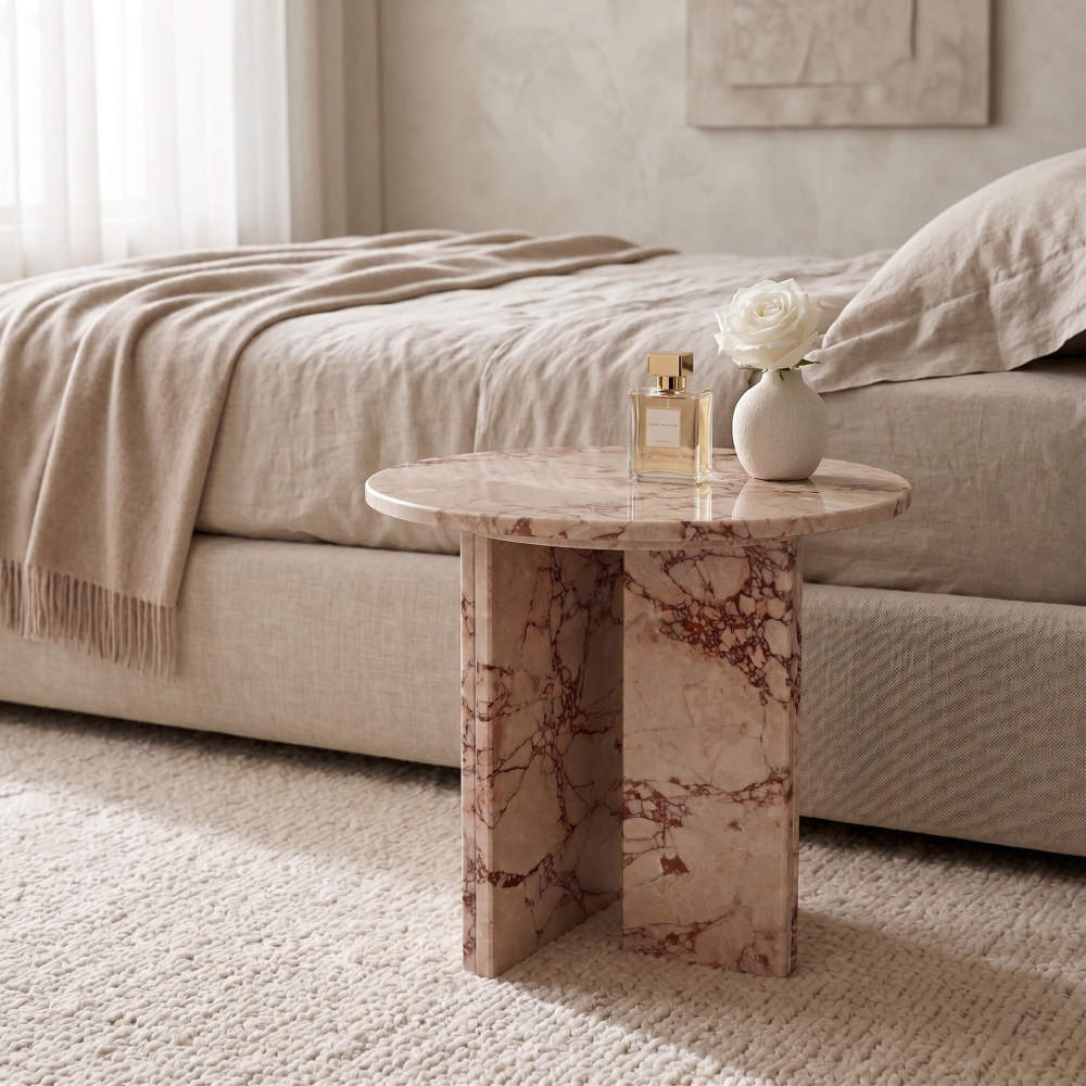 Diamond Pink marble side table used as a bedside nightstand in a Scandinavian minimalist bedroom — linen bedding, chunky knit rug, white rose in ceramic vase, gold-cap perfume bottle on polished stone surface, dual-panel cross base.