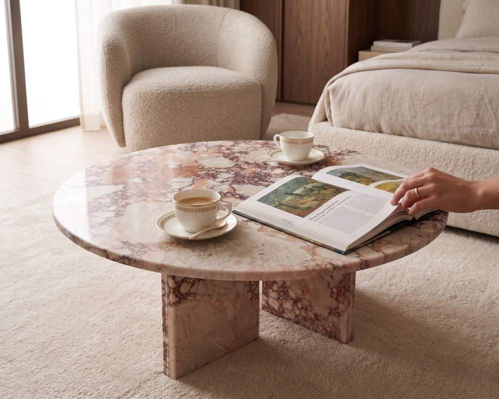 A round pink marble accent table paired with a white bouclé armchair in a sun-drenched Japandi bedroom, highlighting its organic texture.