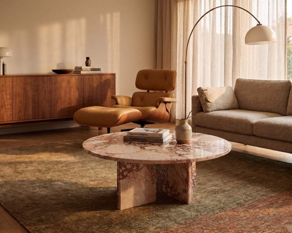 A low-profile pink marble coffee table illuminated by golden hour light in a mid-century modern lounge with walnut furniture and an Eames chair.