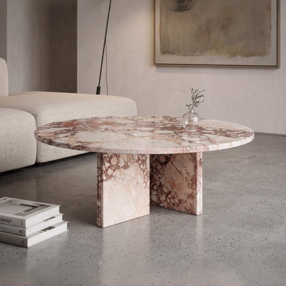 A brecciated pink natural marble coffee table anchoring a minimalist wabi-sabi living space with concrete floors and soft modular seating.