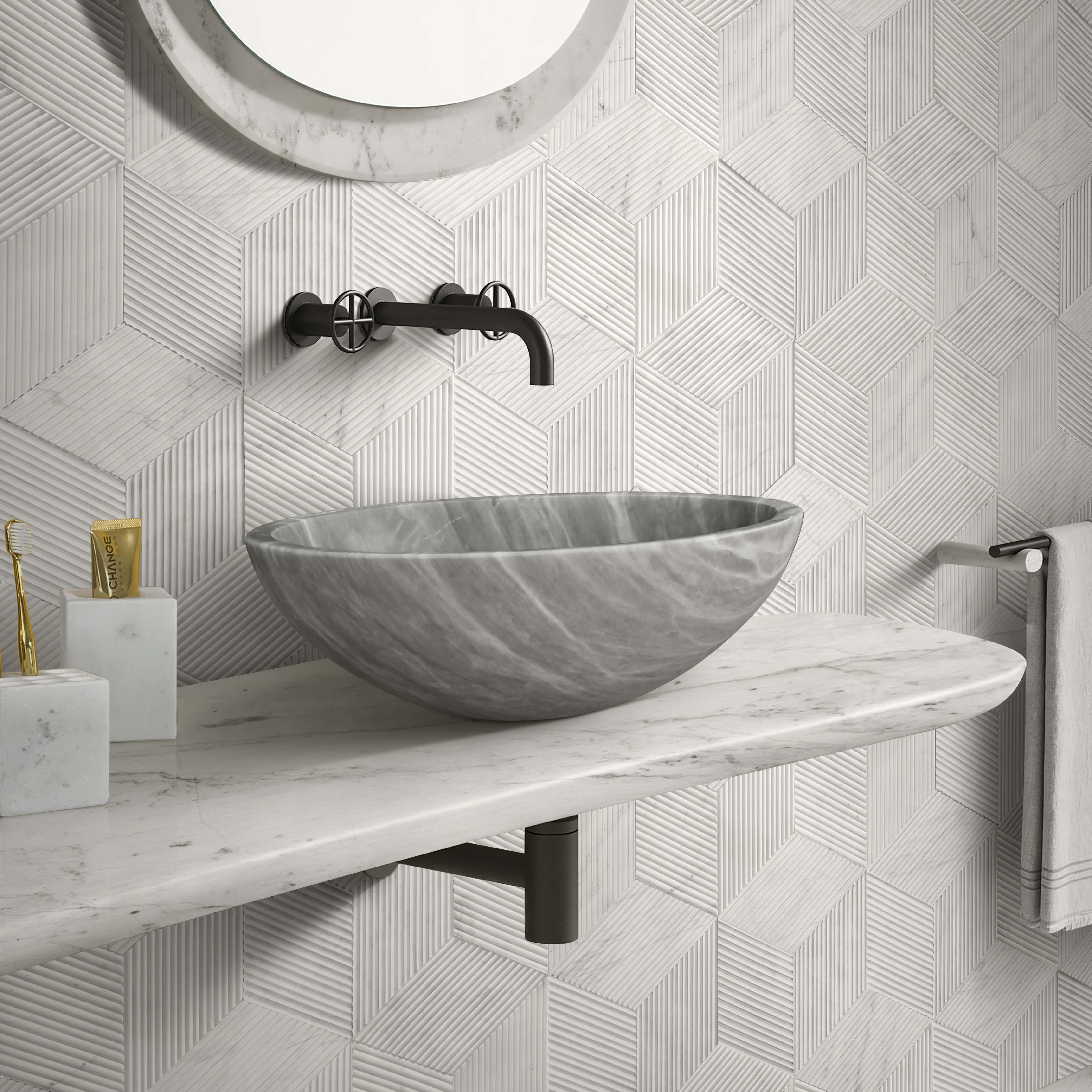Carrara gray marble round vessel sink on a Carrara white marble floating shelf in a boutique hotel-style powder room, against Snow Mountain White hexagon OL-textured finished marble wall tiles with three-directional linear grooves creating a dimensional cube illusion, paired with two pieces from the Snow Mountain White honed-finished marble bathroom accessories set including toothbrush holder and apothecary container, matte black wall-mounted faucet with cross handles, marble-framed round mirror.