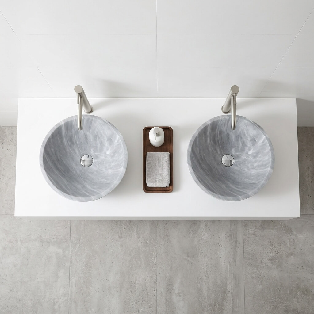 Overhead top-down view of two Carrara gray marble round vessel sinks on a clean white floating countertop with brushed nickel wall-mounted faucets, dark walnut tray with white dispenser and folded gray towel between sinks, concrete-look gray tile flooring, contemporary minimalist bathroom.